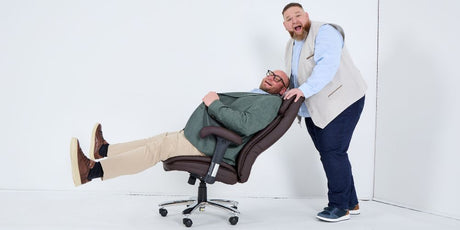 Ein Mann sitzt auf einem Bürostuhl mit 150 kg Belastbarkeit und wird von einem anderen Mann angeschoben. Beide lachen.